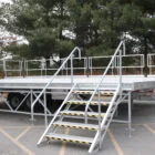 Metal platform trailer with stairs and handrails in a parking lot.