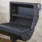 Outdoor grill with open lid and shelf against brick wall.