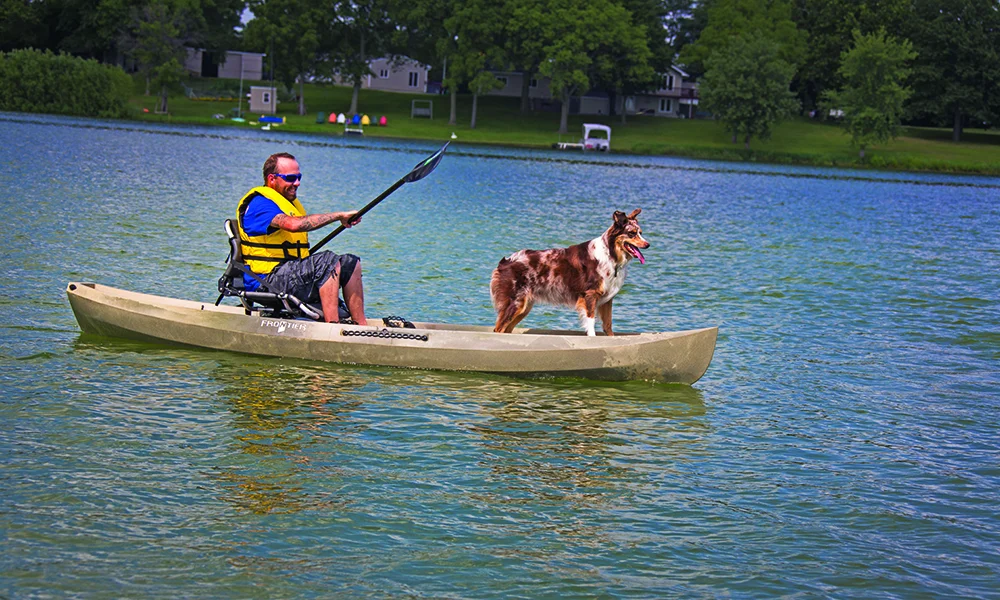 Man paddling kayak on lake with dog standing in front.