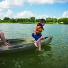 A young boy in a life jacket jumps into a kayak on calm water.