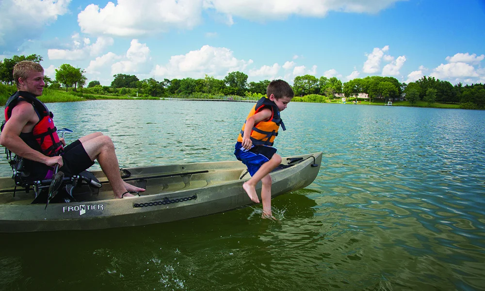 A young boy in a life jacket jumps into a kayak on calm water.