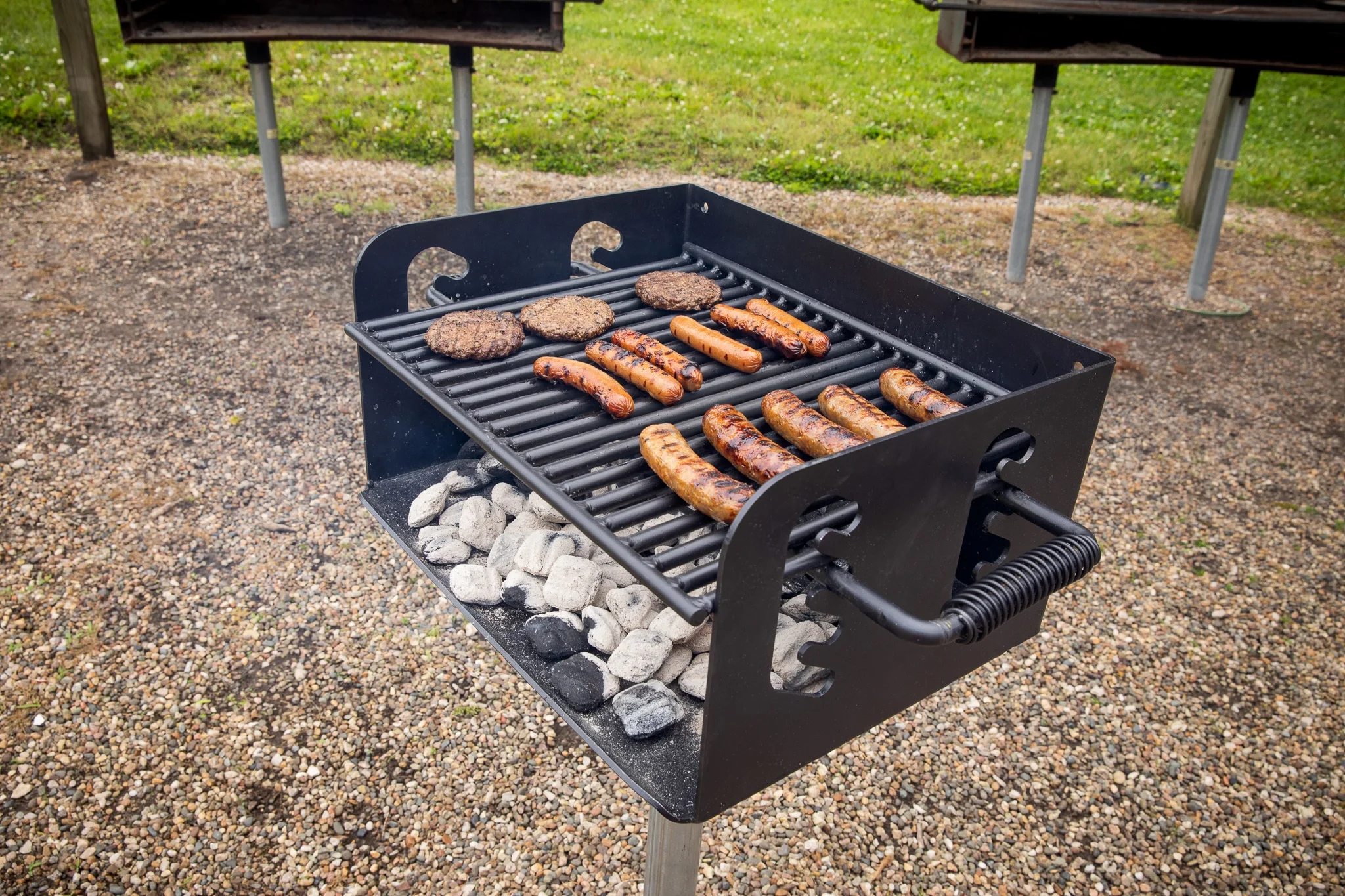 Outdoor grill cooking burgers and sausages on a stone-lined base.