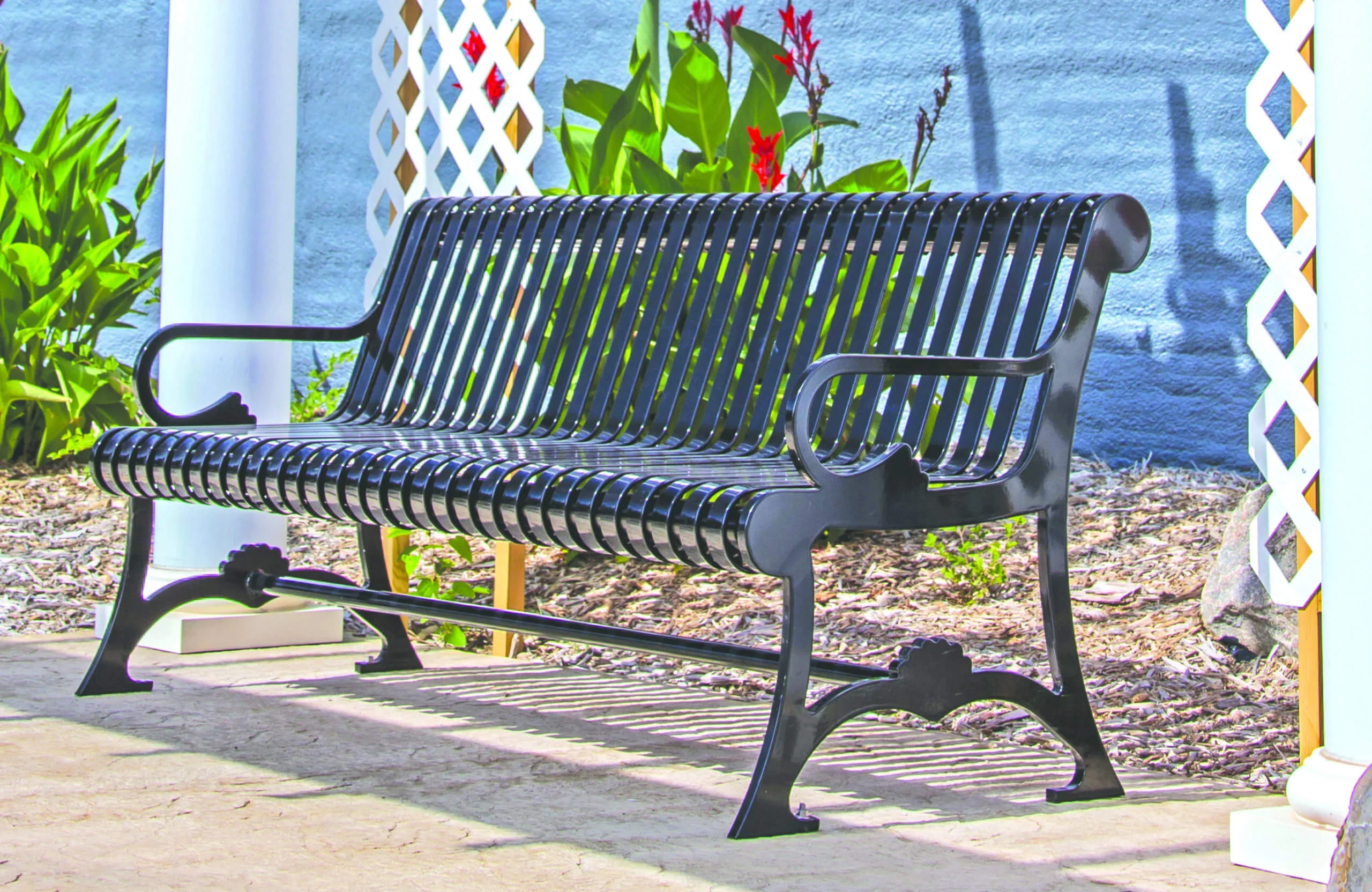 Black metal park bench in front of a blue wall and plants.