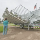 A woman stands next to a large metal trailer with an extended ramp. The trailer is parked on a dirt road near a grassy area and has an American flag flying from its back end.