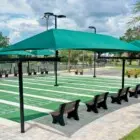 Outdoor shuffleboard court with green turf and black benches under teal umbrellas.