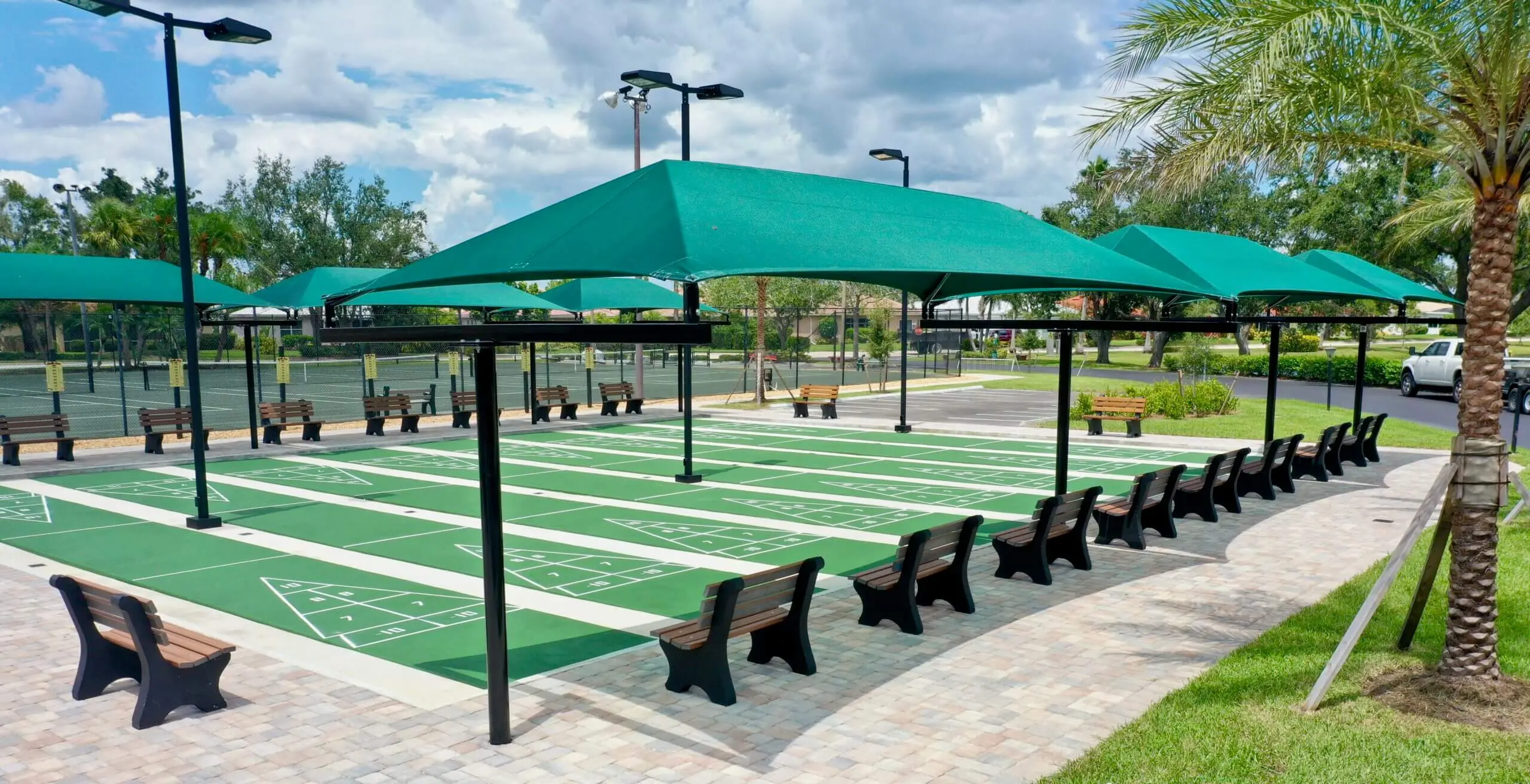 Outdoor shuffleboard court with green turf and black benches under teal umbrellas.