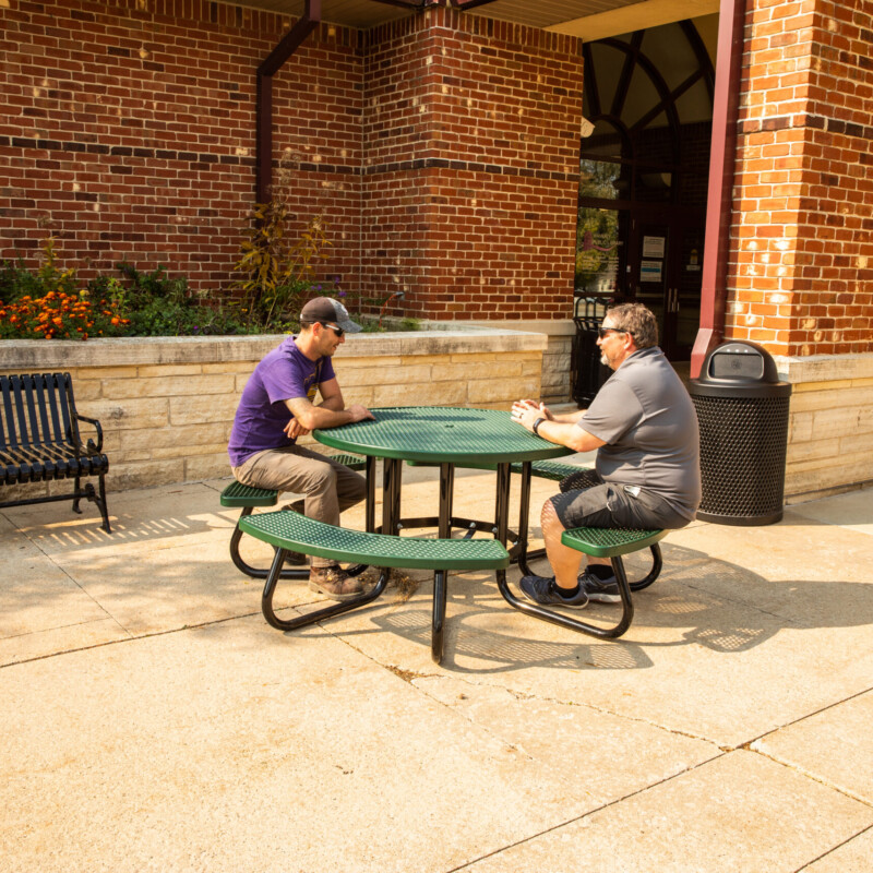 Two people sitting at a green picnic table outside a brick building.