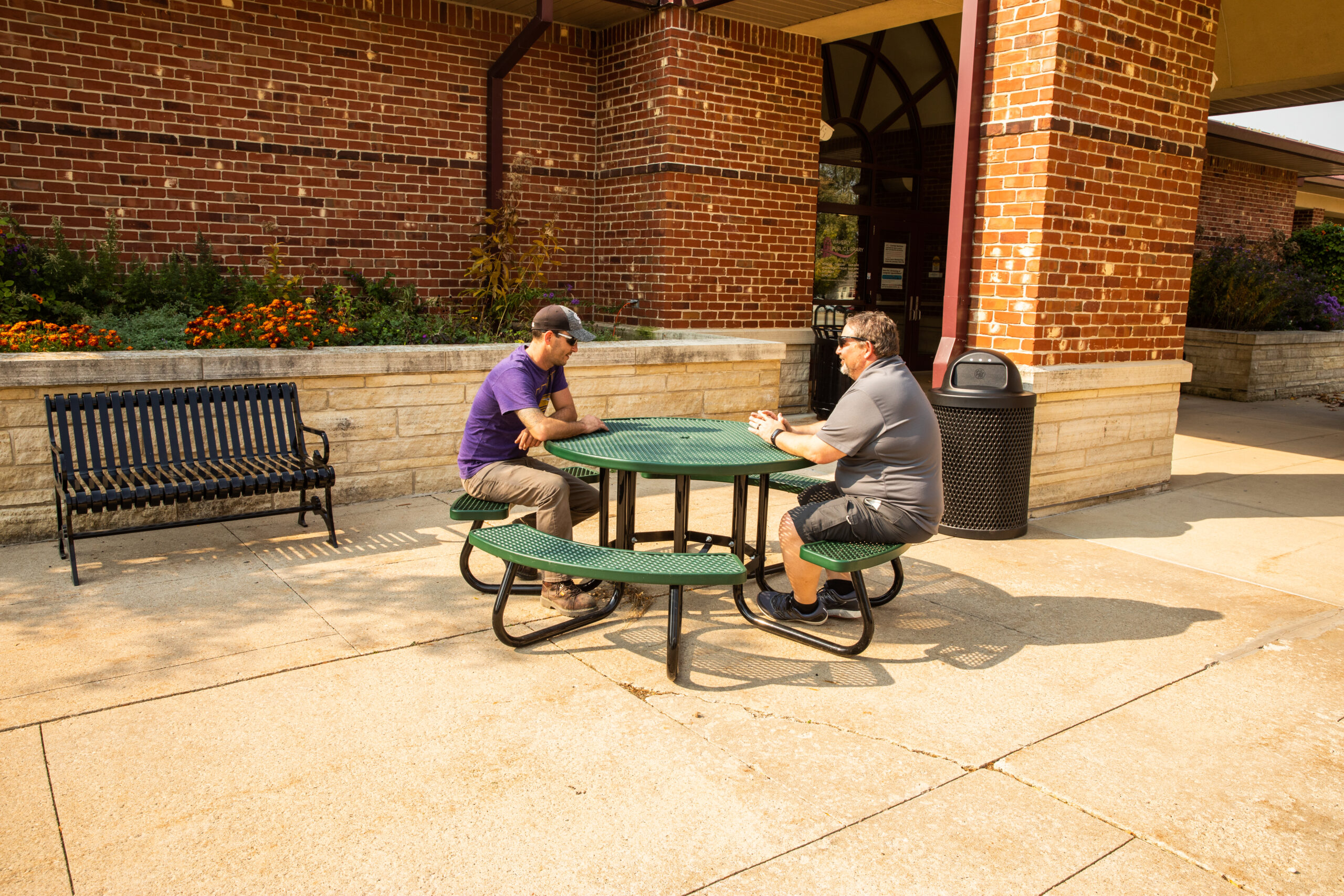 Two people sitting at a green picnic table outside a brick building.